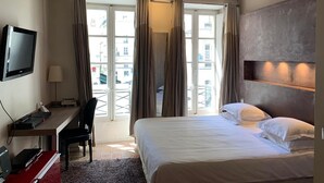 A hotel room with a bed, a desk with a computer, a TV, and a view of the city through large windows. - Le Marais Sicile (Paris)