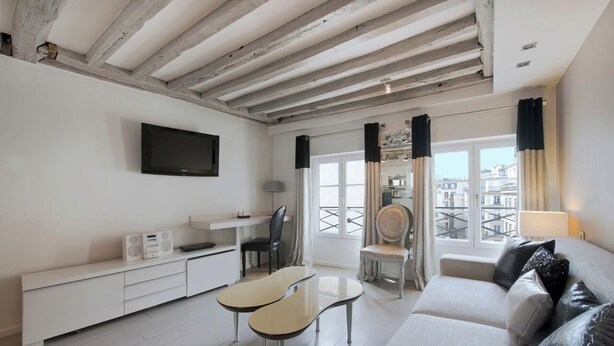 A modern living room with a white sofa, a TV mounted on the wall, a small dining table, and a balcony with a view of the city. - Le Marais Sicile (Paris)