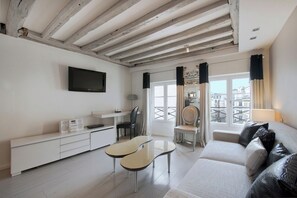 Un salon moderne avec un canapé blanc, un téléviseur fixé au mur, une petite table à manger et un balcon offrant une vue sur la ville.