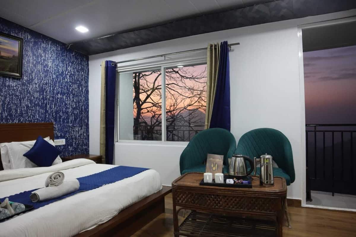 A hotel room with a bed, a window with a view of trees and a sunset, a small table with a kettle and cups, and a chair.