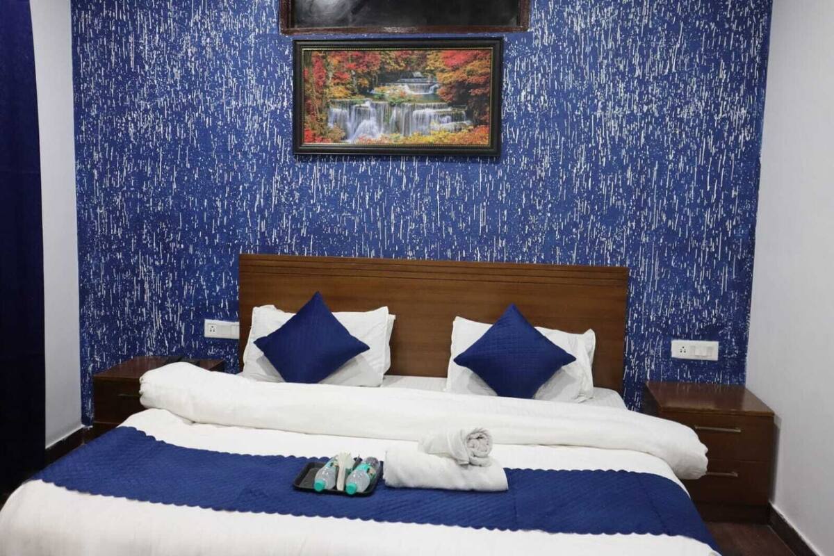 A neatly made bed with white and blue bedding, two pillows, and a blue blanket. There are two bedside tables with lamps, a framed picture above the bed, and a small tray with toiletries on the bed.