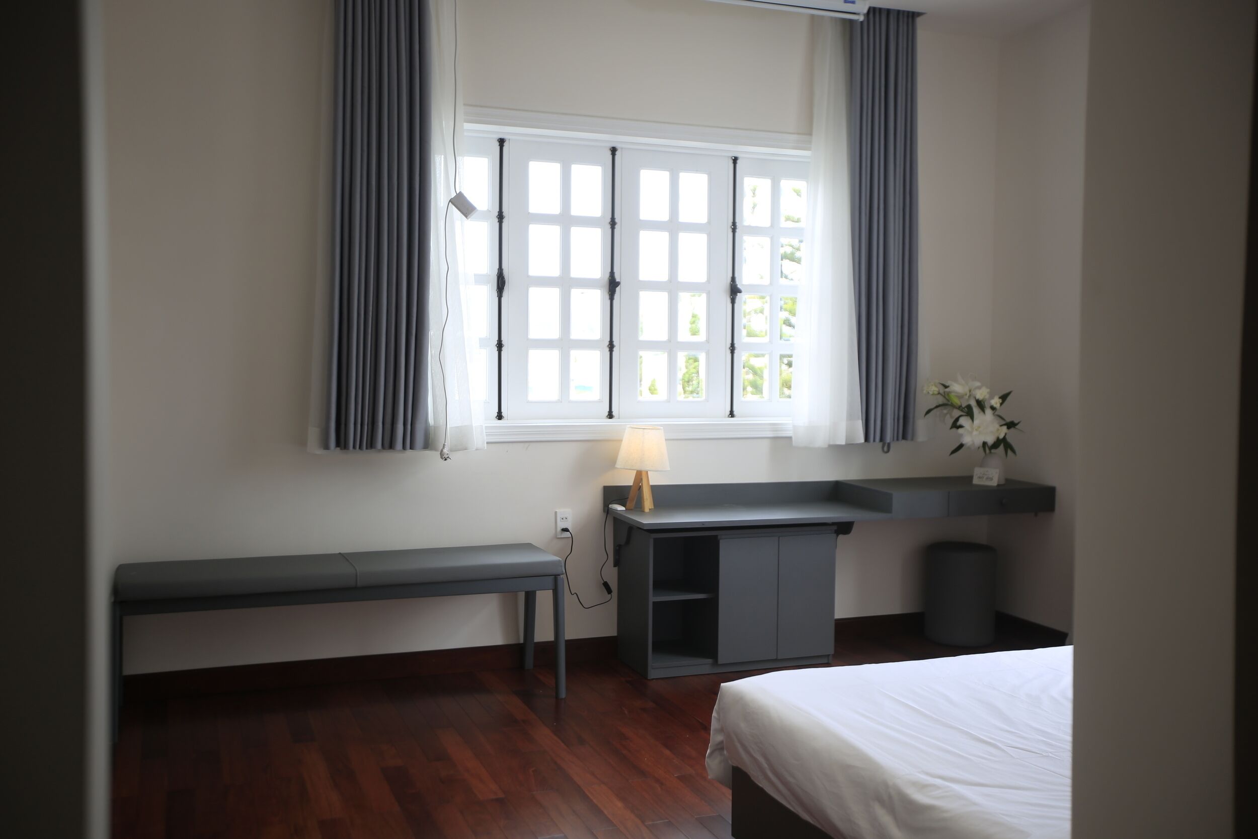 A bedroom with a bed, a desk, a bench, a window with curtains, and a small plant.