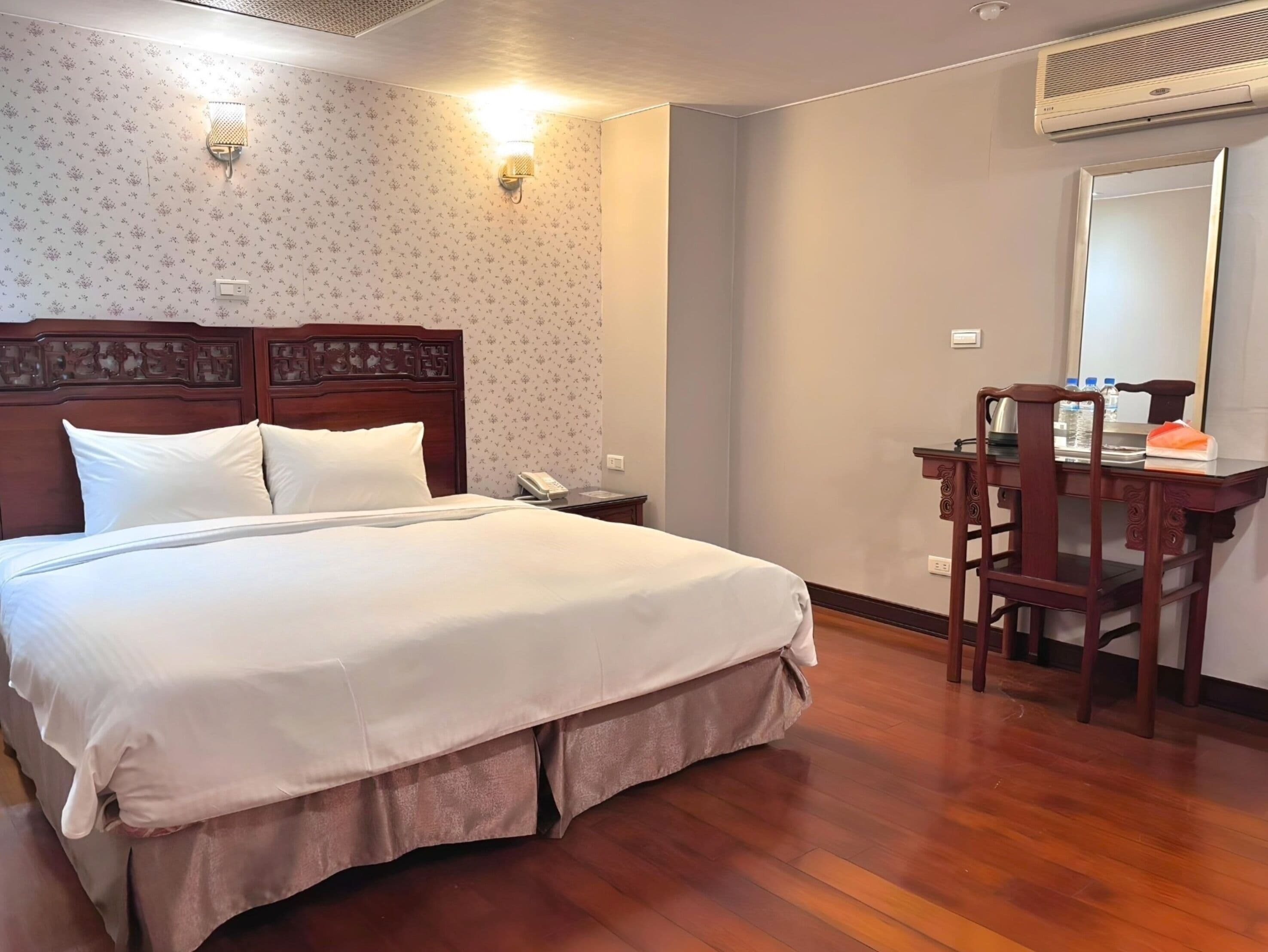 A neatly made bed with a wooden headboard, a desk with a chair, and a wall-mounted air conditioner.