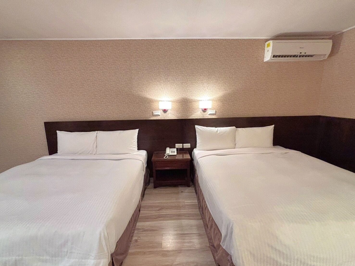 Quarto de hotel com duas camas de solteiro, uma mesinha de cabeceira com telefone e um aparelho de ar condicionado na parede.