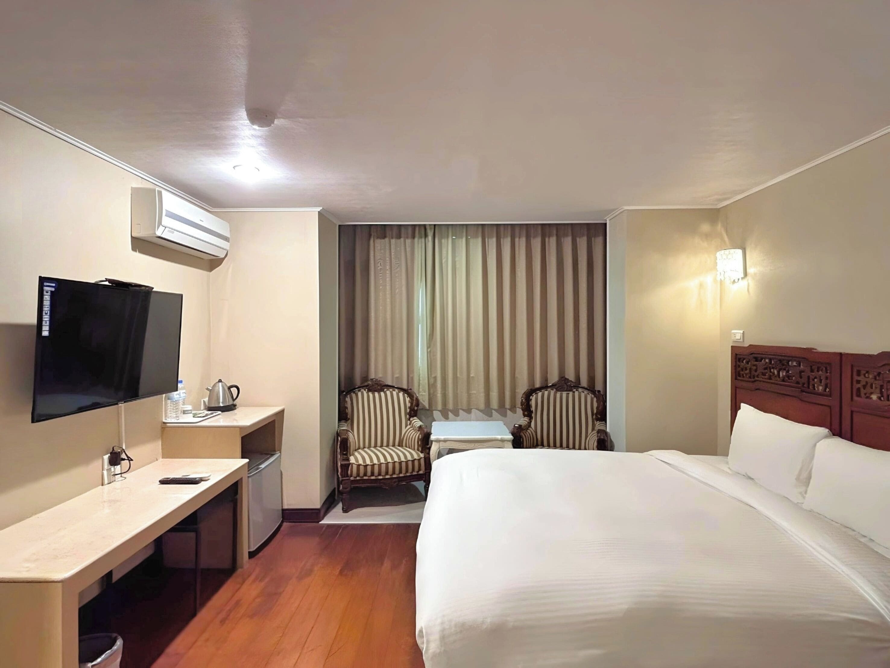 A hotel room with a bed, a desk, a television, two chairs, a window with curtains, and a wall-mounted light.
