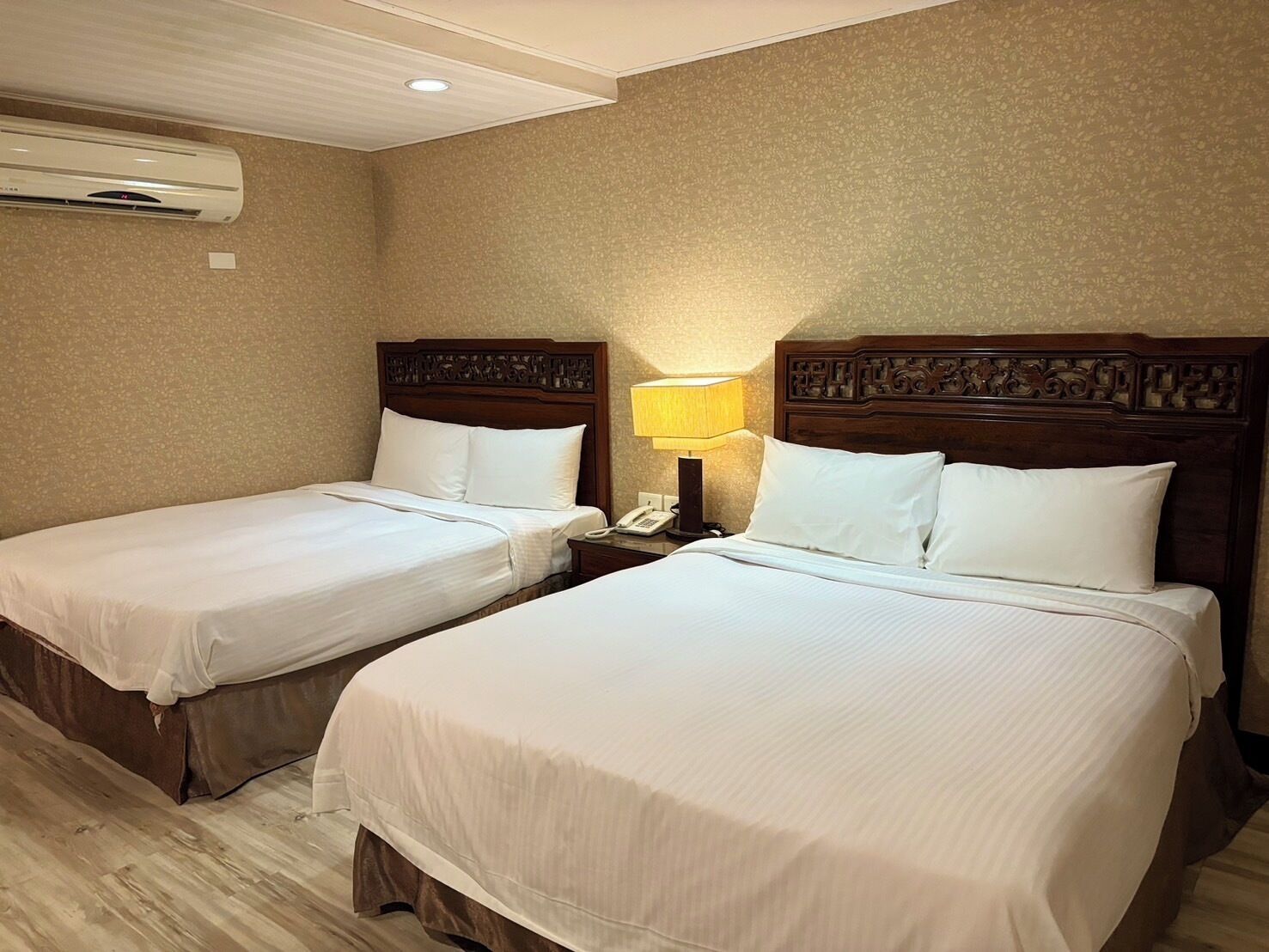 Habitación de hotel con dos camas individuales, cabecero de madera, lámpara de noche y aire acondicionado en la pared.