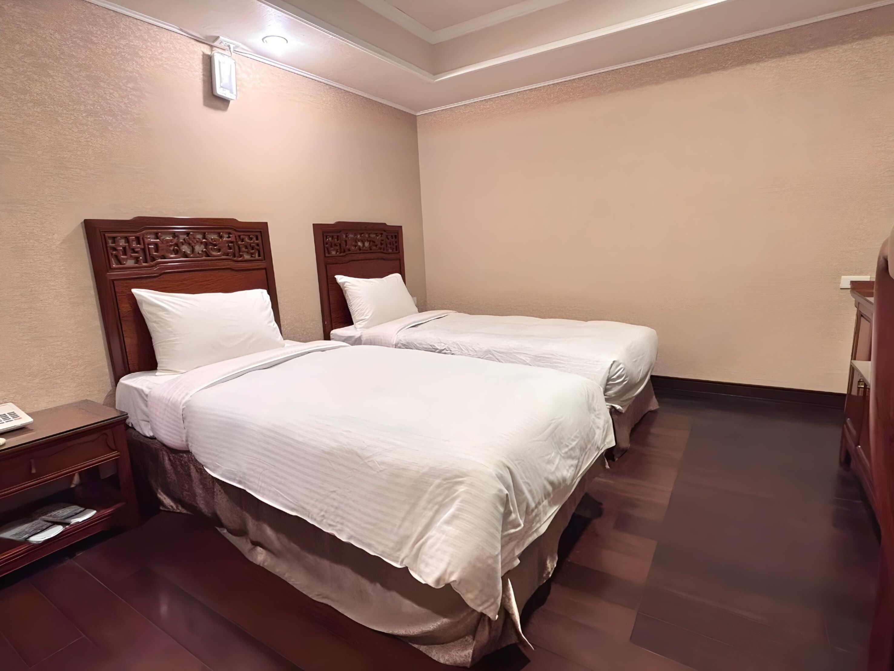 A hotel room with two beds, wooden headboards, a nightstand with a lamp, and a small table.