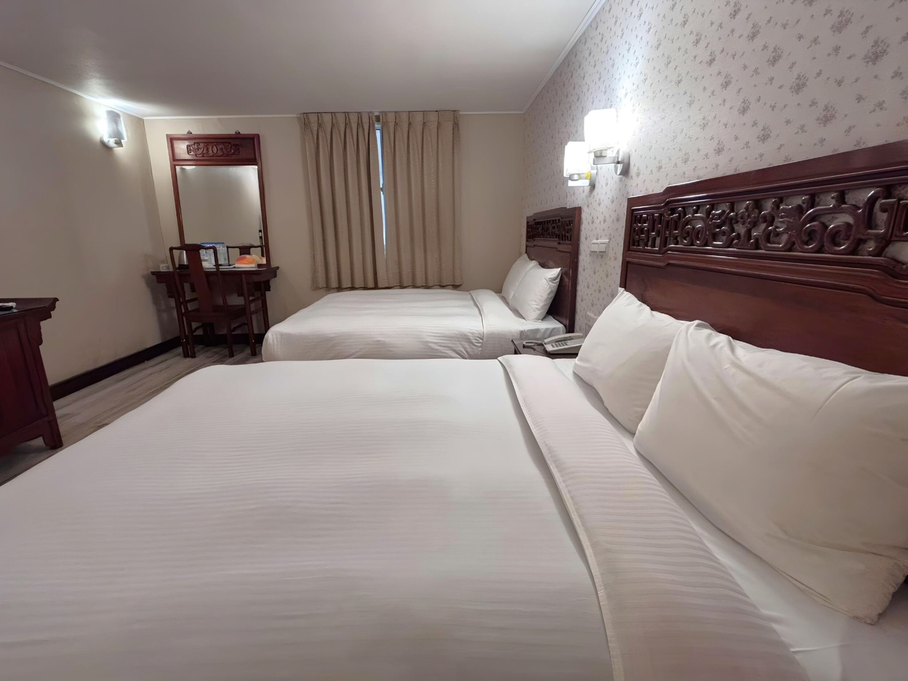 A hotel room with two beds, a desk, a chair, a mirror, and a window with curtains.
