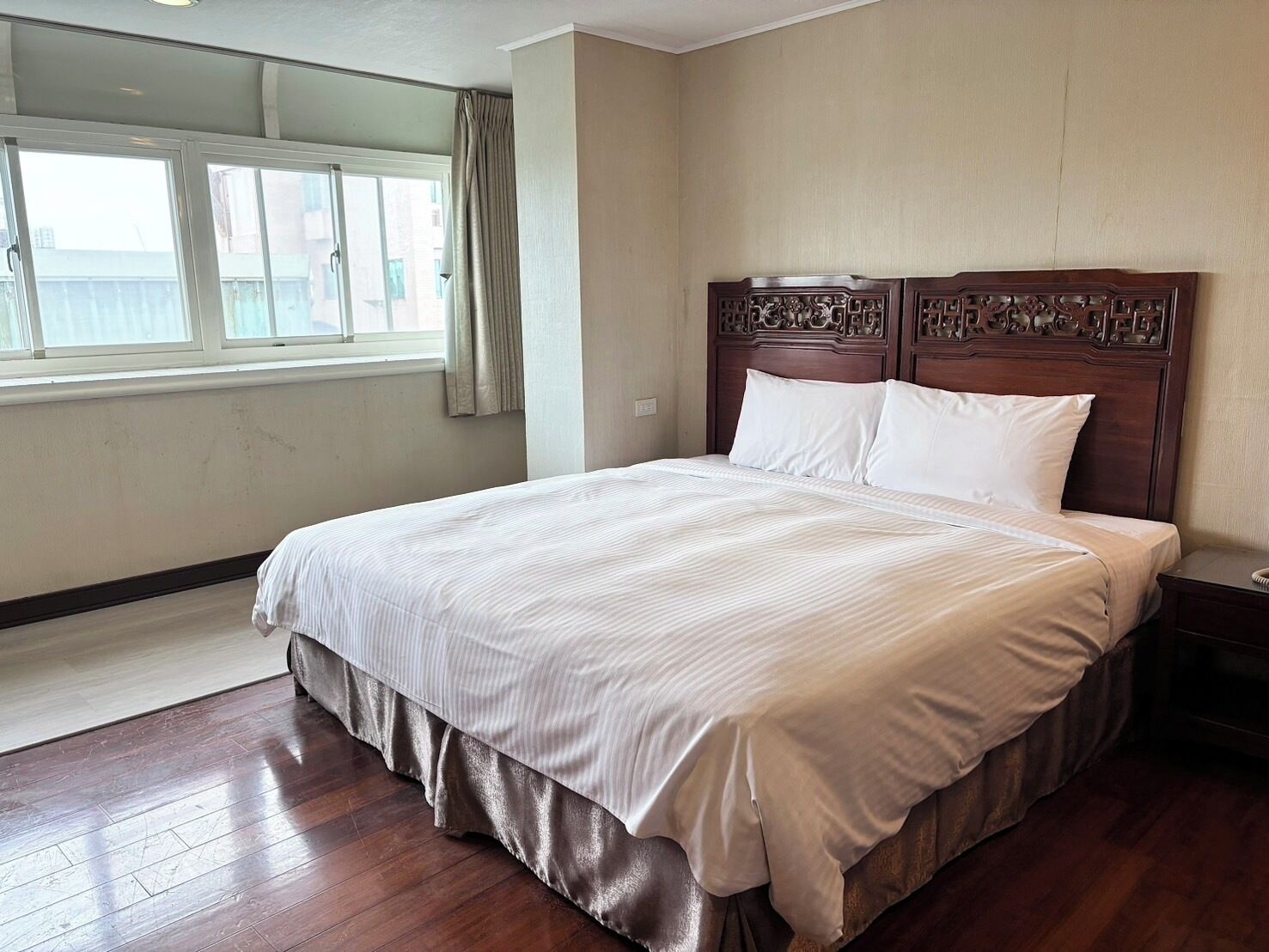 A hotel room with a large bed, a wooden headboard, a window with curtains, and a wooden floor.