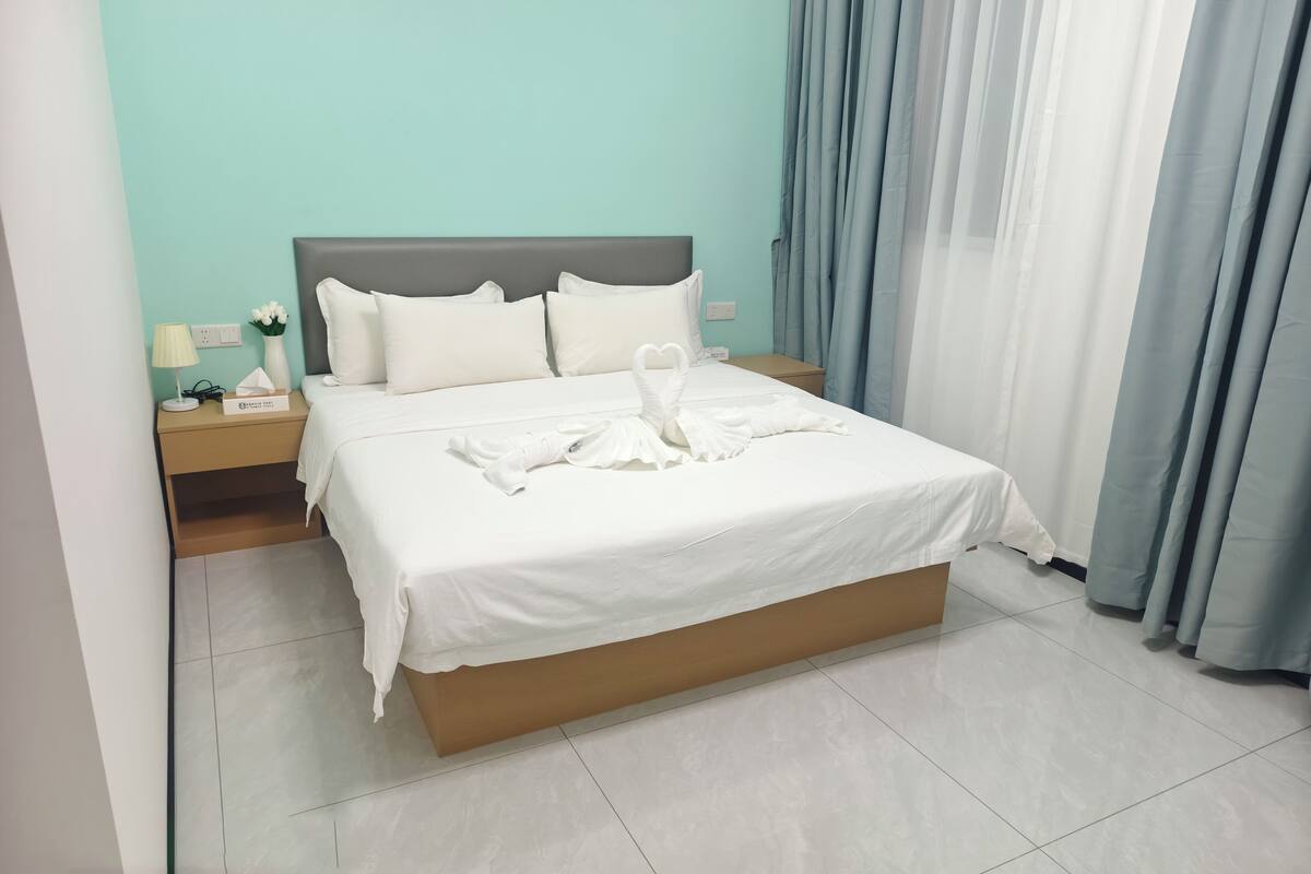 A neatly made bed with white linens and a swan-shaped towel arrangement. There is a nightstand with a lamp and a vase of flowers. The room has a teal wall and light blue curtains.