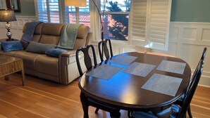 Un salon avec un canapé en cuir marron, une table basse en bois et une table à manger avec quatre chaises. Deux lampes suspendues sont placées au-dessus de la table à manger.