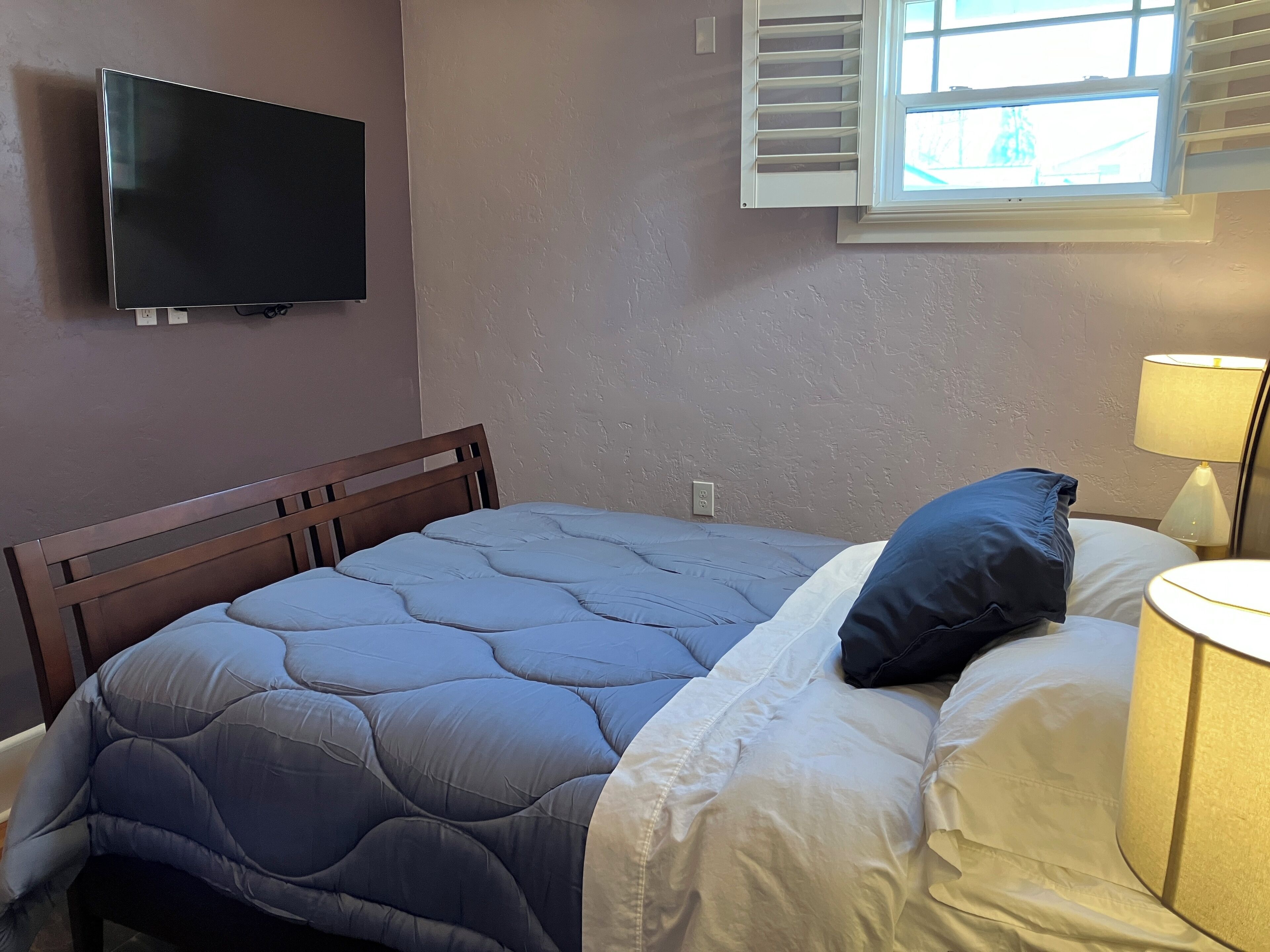 Un dormitorio con cama, televisión montada en la pared, ventana con contraventanas y lámpara de noche.