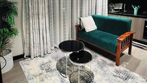 Un salon avec un canapé vert, deux petites tables rondes noires, un tapis à motifs et une grande fenêtre ornée de rideaux légers.