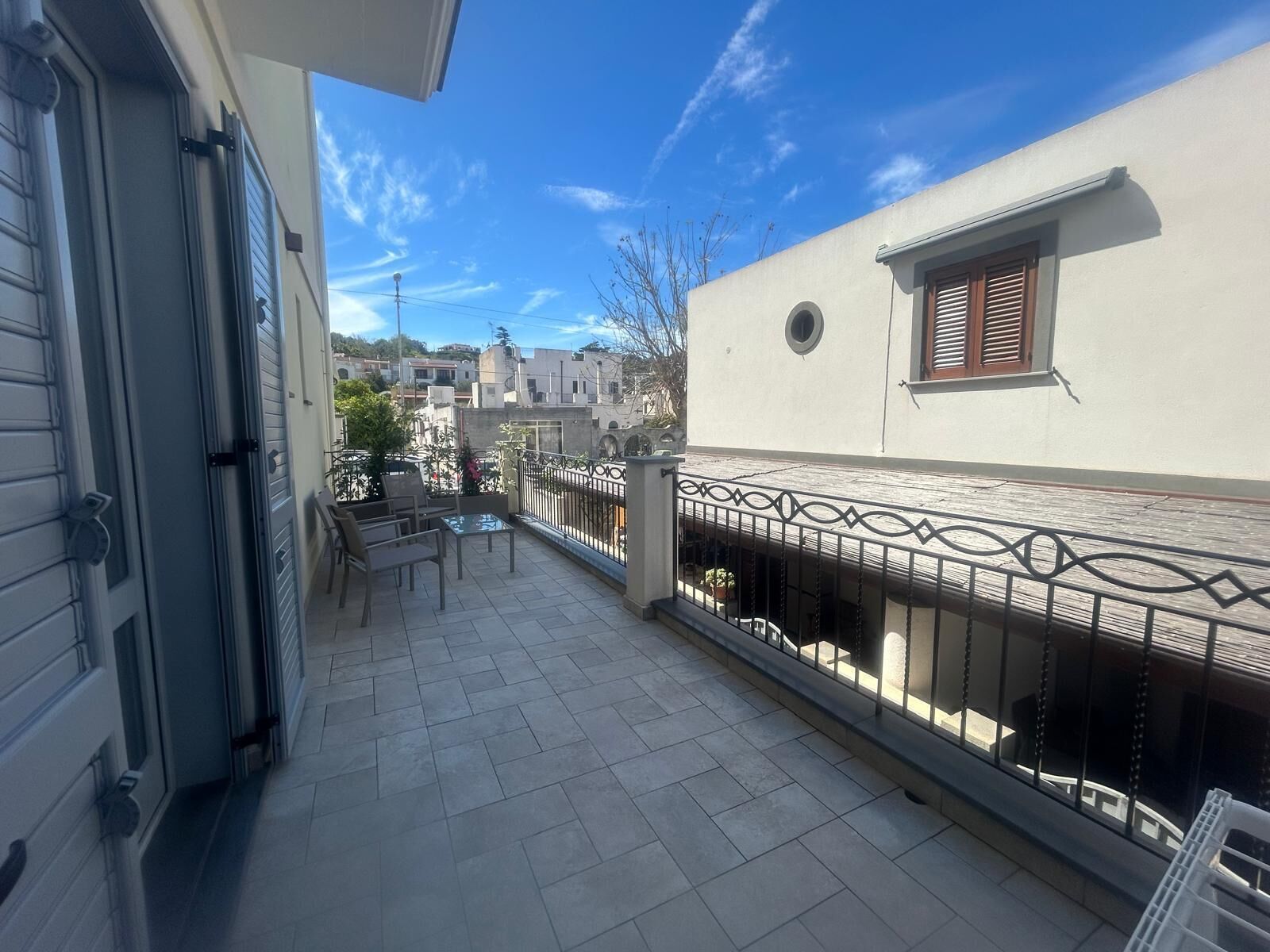 A balcony with a tiled floor, a small table, and chairs. There is a railing with decorative elements.