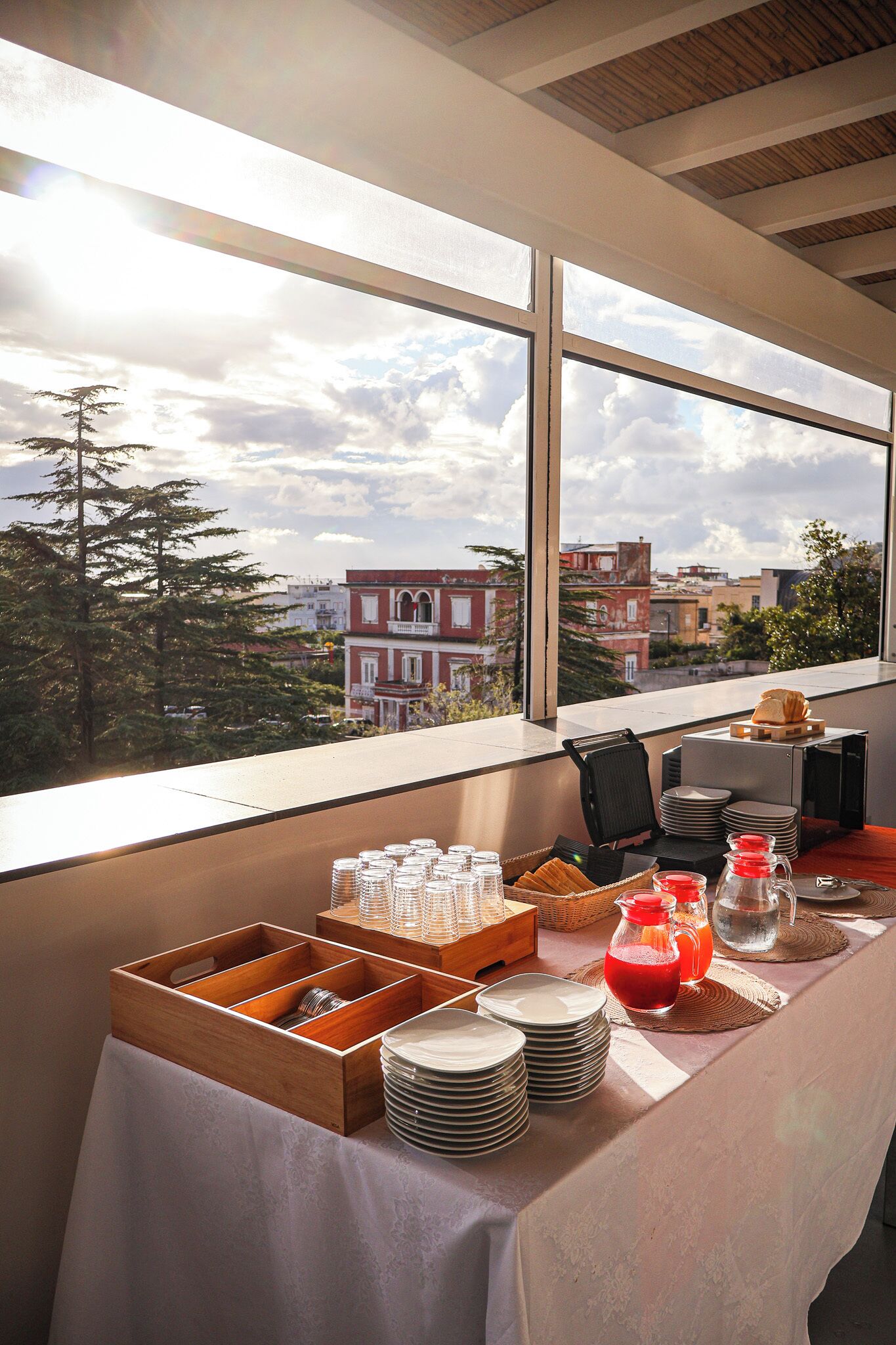 Mesa posta para o café da manhã, com pratos, copos e jarra de suco, exibindo vista para edifícios e árvores através de janelas amplas.