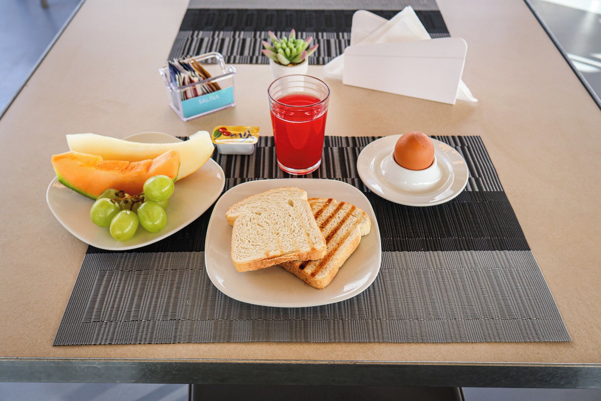 A table with a plate of sliced bread, a plate of melon and grapes, a glass of red juice, and a small dish with an egg.