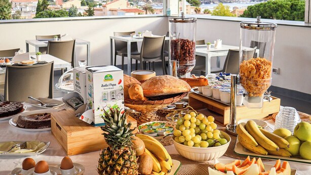 Un bufé de desayuno con diversas frutas, pan y bebidas, en un salón con vistas a la ciudad.