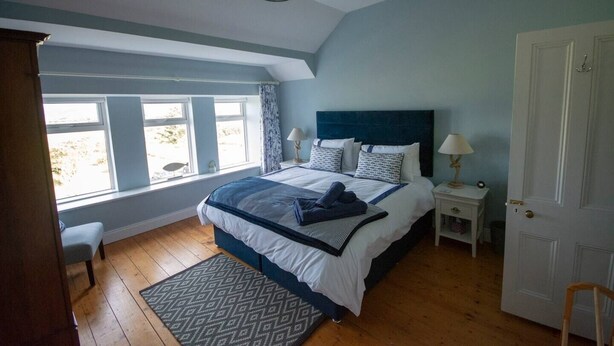 A bedroom with a large bed, two bedside tables, a window with curtains, a door, and a wooden floor. - Paddy's Atlantic View! Home for 8 in Waterville! (Waterville)