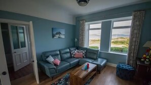 A living room with a blue sofa, a wooden coffee table, and a view of the outdoors through large windows. - Paddy's Atlantic View! Home for 8 in Waterville! (Waterville)