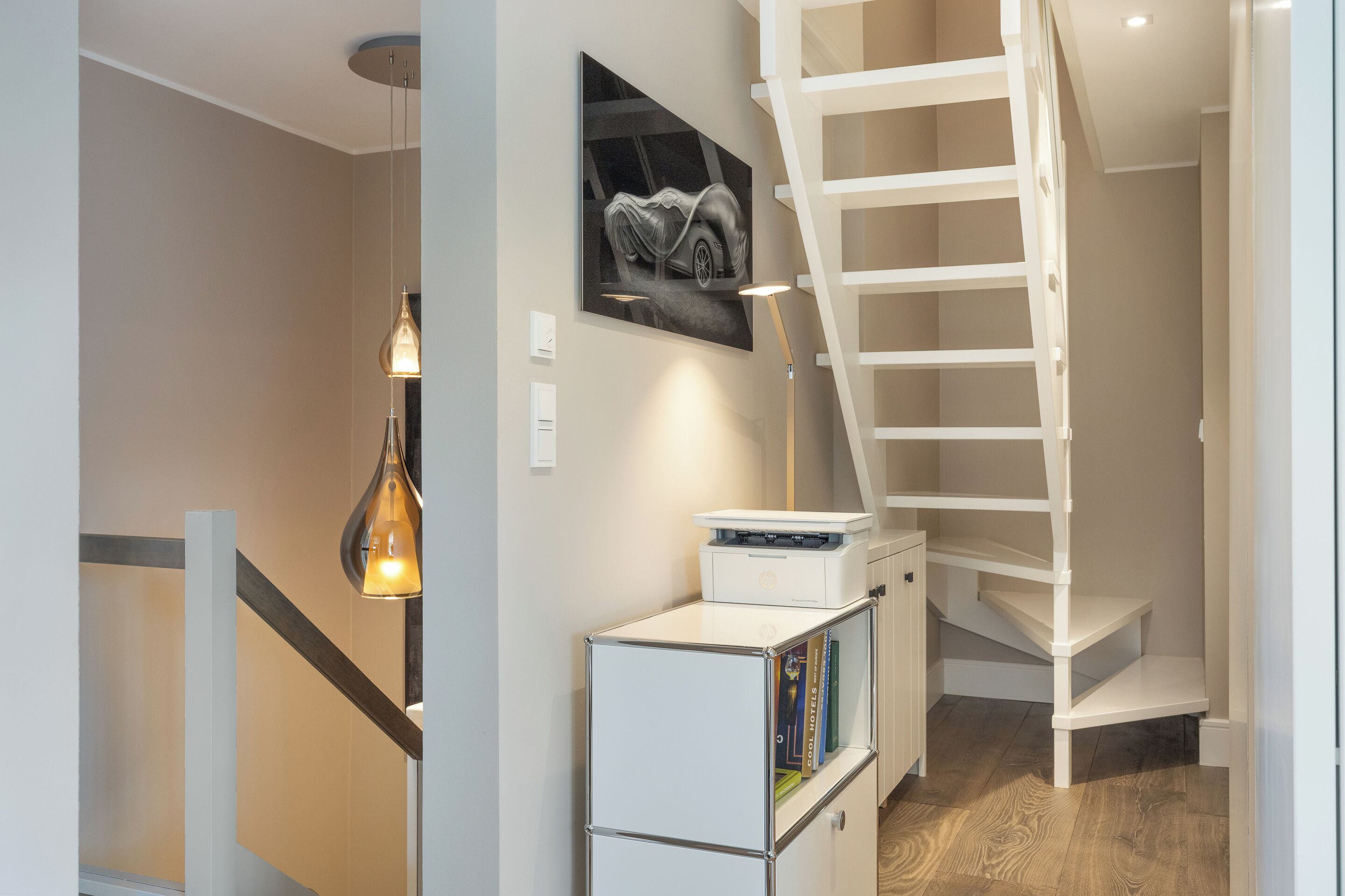 A modern interior with a staircase, a wall-mounted artwork, a desk with a printer, and a hanging light fixture.