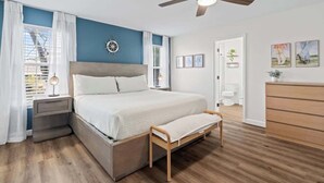 A bedroom with a bed, bedside table, bench, ceiling fan, and a view of the bathroom.