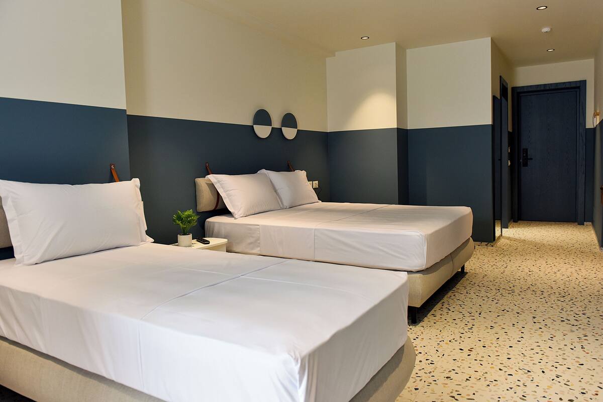 Ein Hotelzimmer mit zwei Betten, einem kleinen Nachttisch mit einer Pflanze und einer Tür zu einem weiteren Raum.