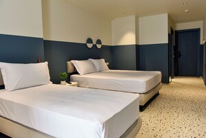 Een hotelkamer met twee bedden, een klein nachtkastje met een plant, en een deur die naar een andere kamer leidt.