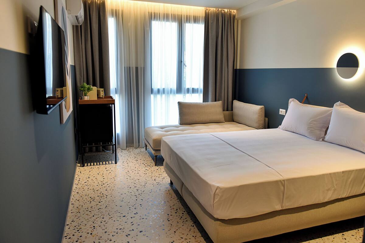 Ein Hotelzimmer mit einem großen Bett, einem Fernseher, einem kleinen Tisch mit Pflanze und einem Fenster mit Vorhängen.