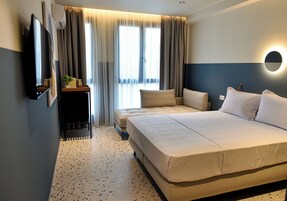 Una habitación de hotel con una cama grande, un televisor, una mesita con una planta y una ventana con cortinas.