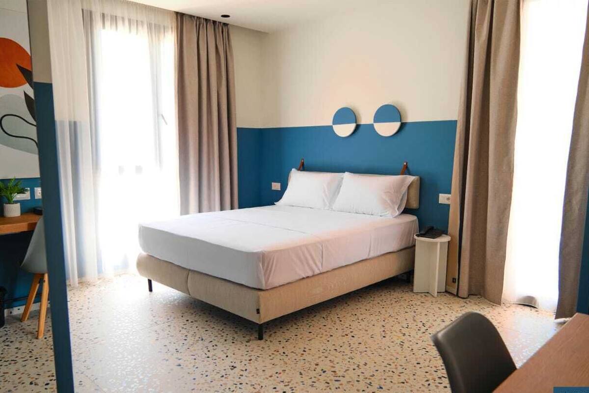 Ein Schlafzimmer mit Bett, Nachttisch, Stuhl, Schreibtisch mit Pflanze und Wand mit abstrakter Kunst.