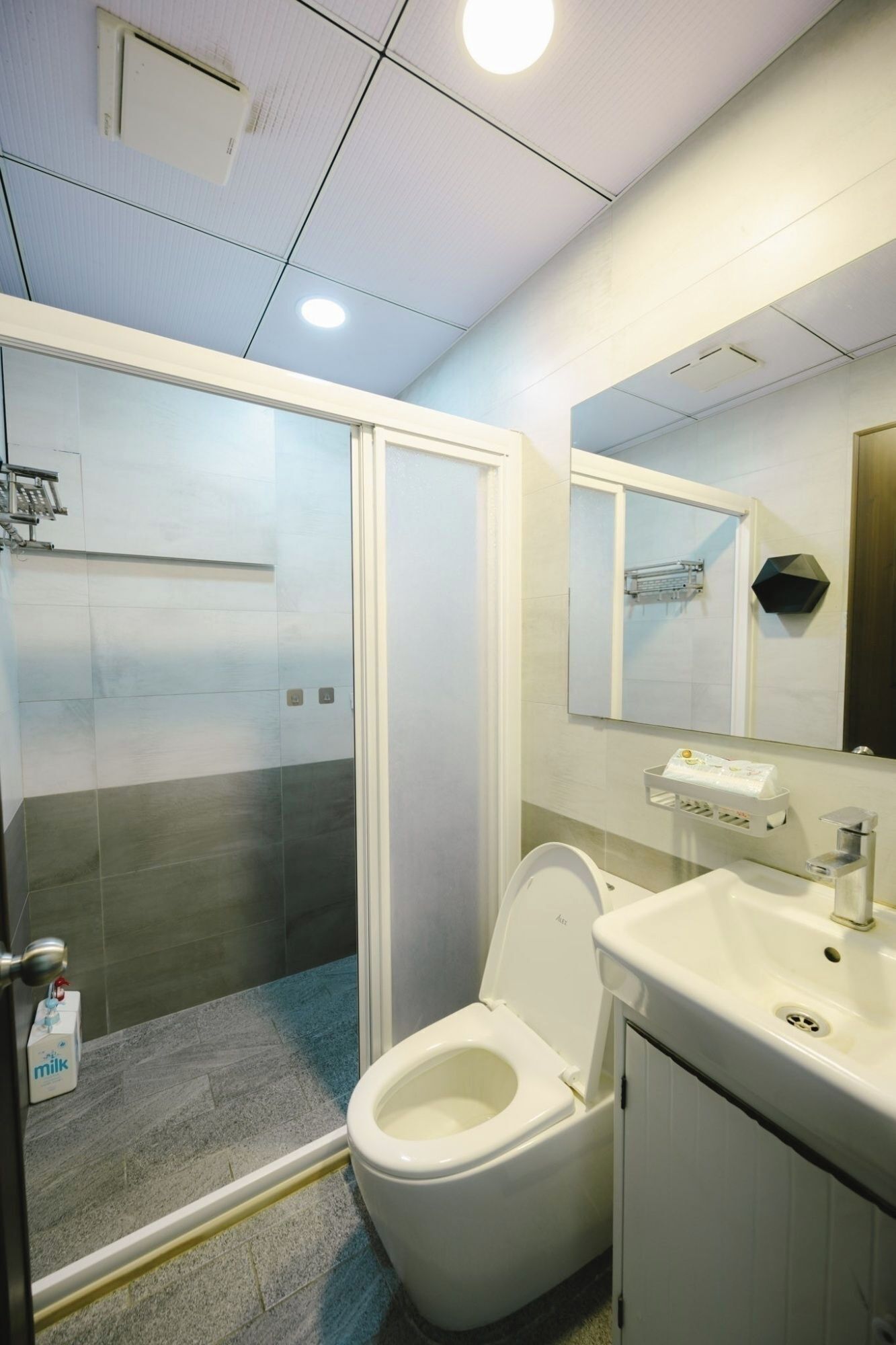Un baño moderno con cabina de ducha de vidrio, inodoro, lavabo con espejo y un pequeño estante con artículos de aseo.