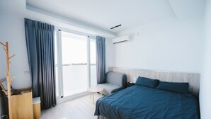 Een moderne hotelkamer met een groot bed, een kleine tafel en een stoel. Er is een raam met gordijnen, een airconditioning en een houten kapstok.