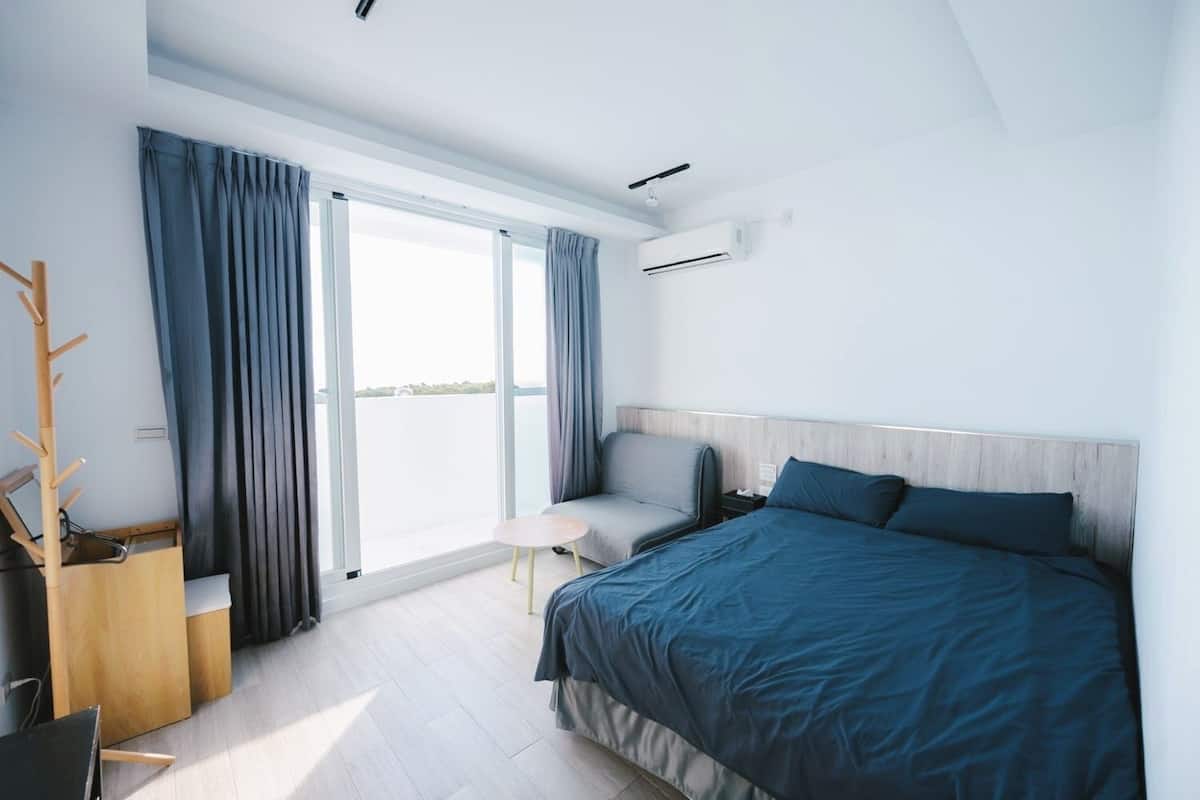 Une chambre d’hôtel moderne avec un grand lit, une petite table et une chaise. On y trouve une fenêtre avec des rideaux, un appareil de climatisation et un porte-manteau en bois.