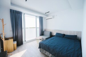 Una habitación de hotel moderna con una cama grande, una mesita pequeña y una silla. Cuenta con una ventana con cortinas, un equipo de aire acondicionado y un perchero de madera.