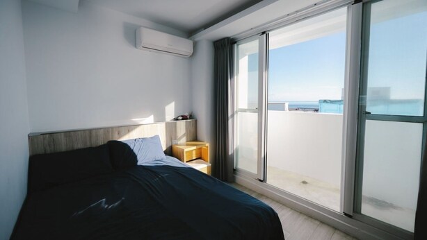 Een moderne hotelkamer met een groot bed, een klein nachtkastje, een airconditioning en een schuifglasdeur naar een balkon met uitzicht op zee.