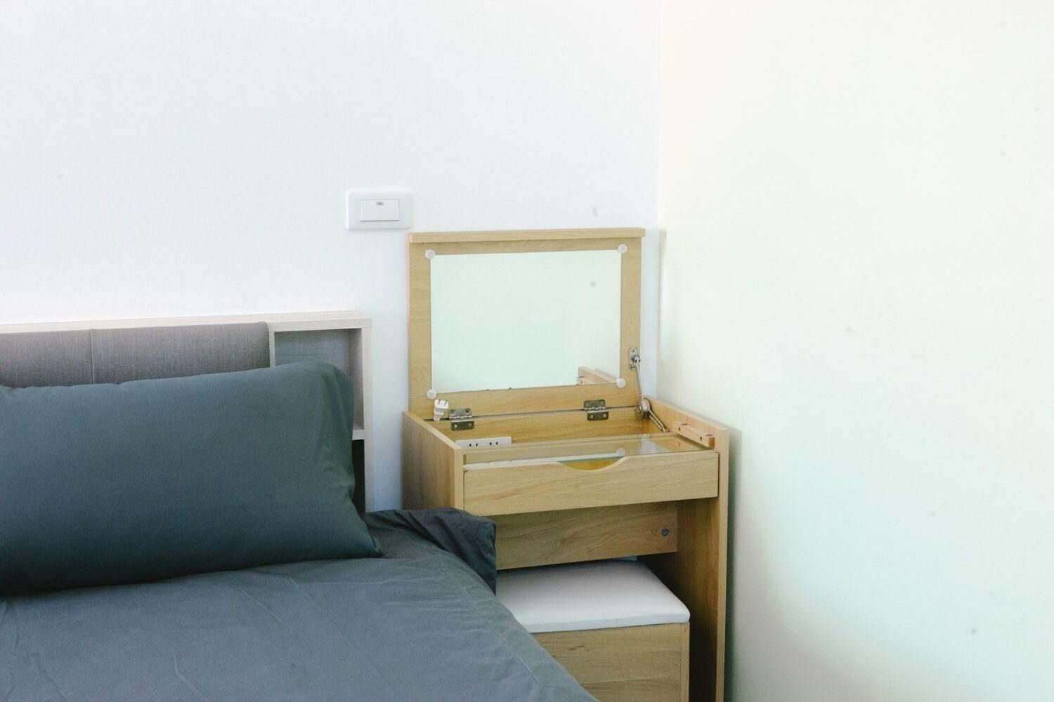 Um quarto pequeno e modesto, com uma cama, uma mesinha de cabeceira de madeira e um espelho.