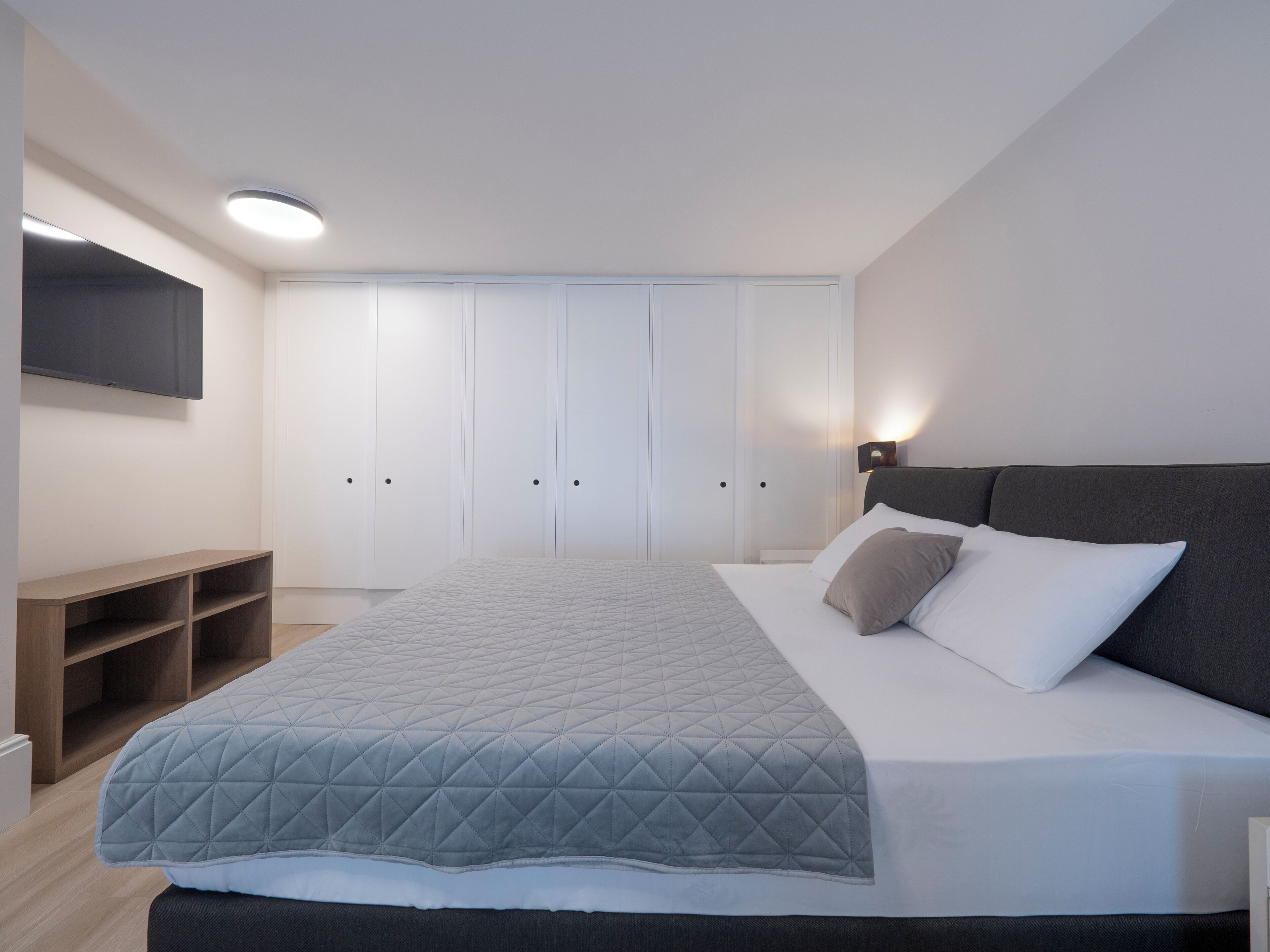 Quarto moderno com cama, televisão fixada na parede e guarda-roupa com várias portas.