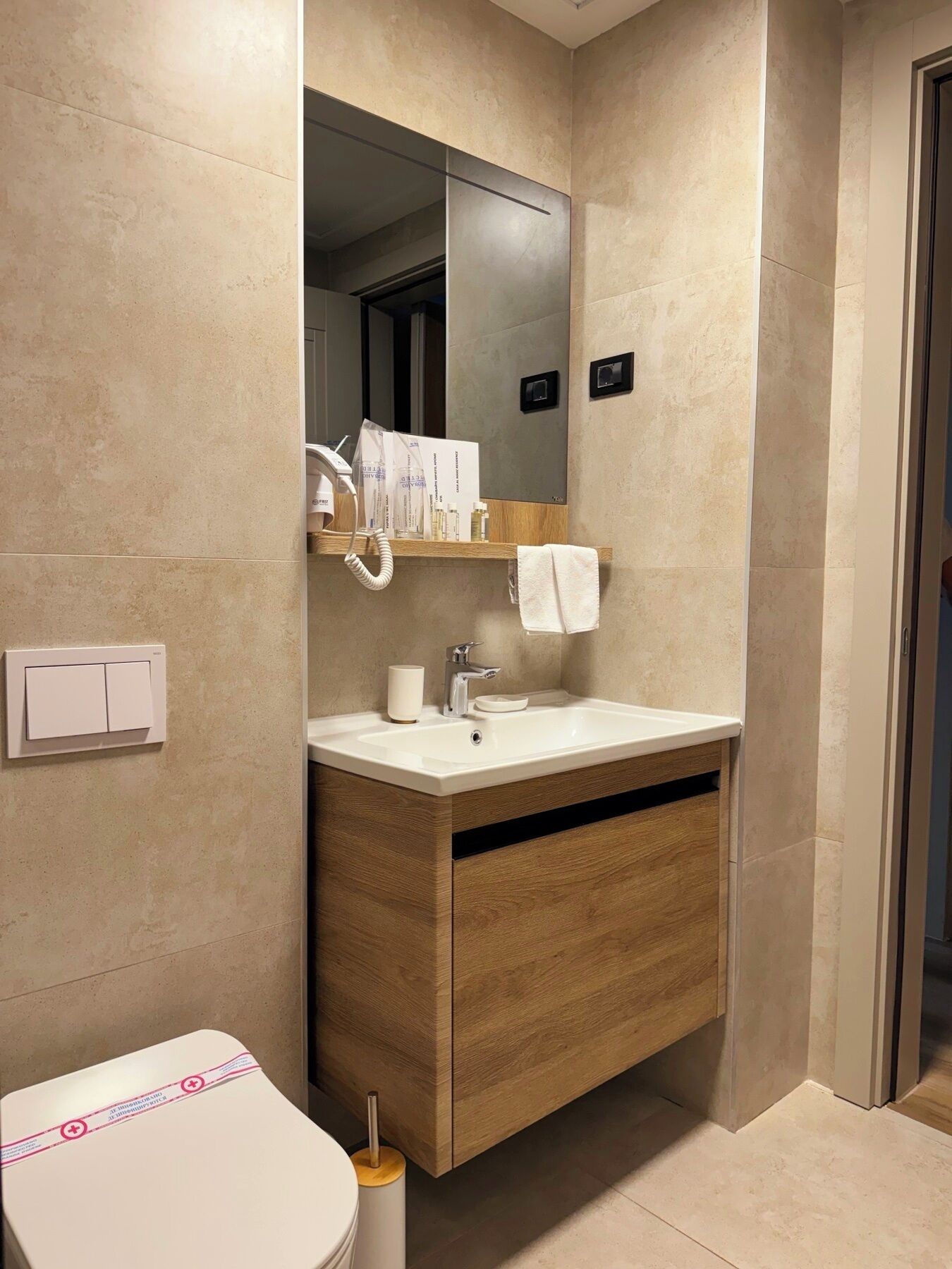 A modern bathroom with a wooden vanity, a white sink, and a mirror above it. There is a towel hanging on the wall and a toilet with a white lid.
