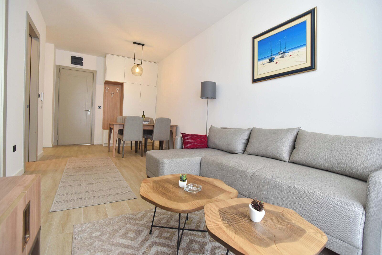 A modern living room with a grey sofa, two wooden coffee tables, a dining area with a table and chairs, and a framed picture on the wall.