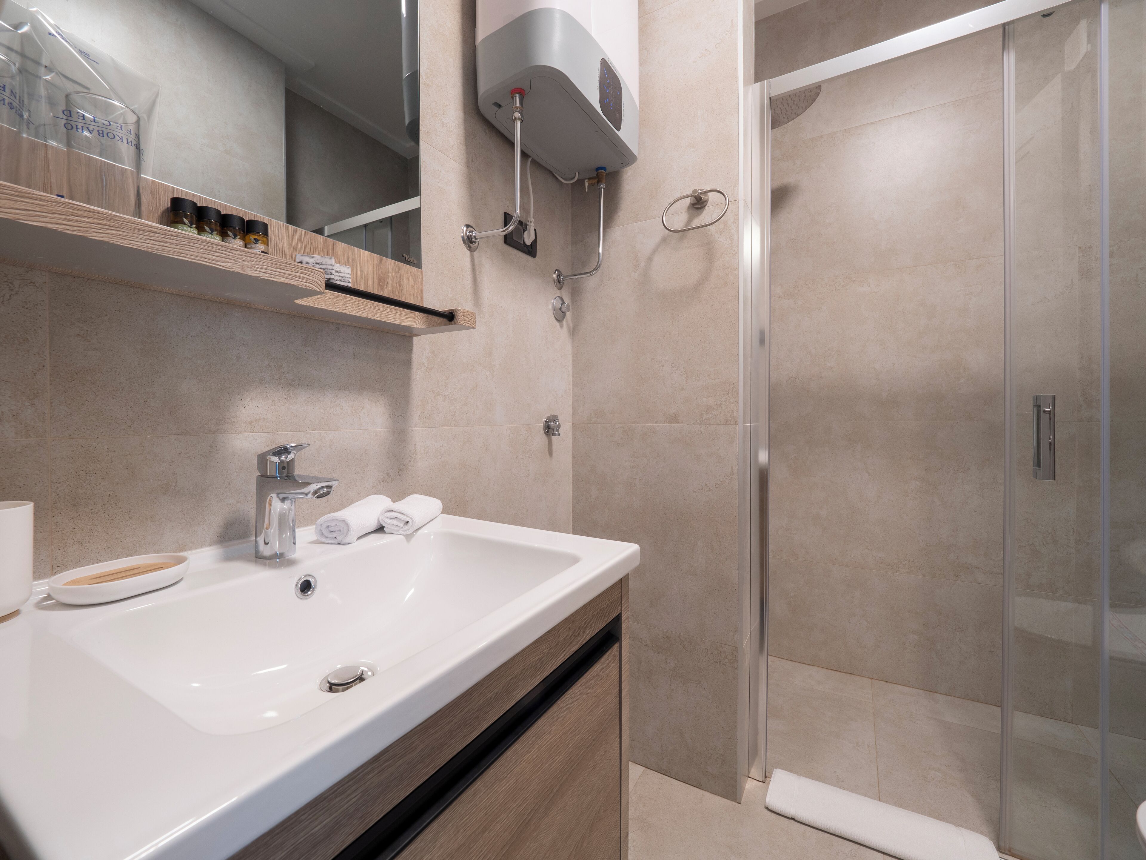 A modern bathroom with a white sink, a glass shower enclosure, and a wooden shelf with toiletries.