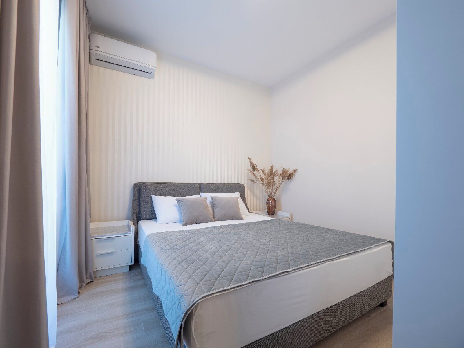Une chambre à coucher moderne, avec un lit, des tables de chevet, un appareil de climatisation et un vase contenant des plantes séchées.