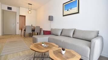Una sala de estar moderna con un sofá gris, dos mesas de centro de madera, una zona de comedor con sillas, una cocina y un cuadro en la pared.
