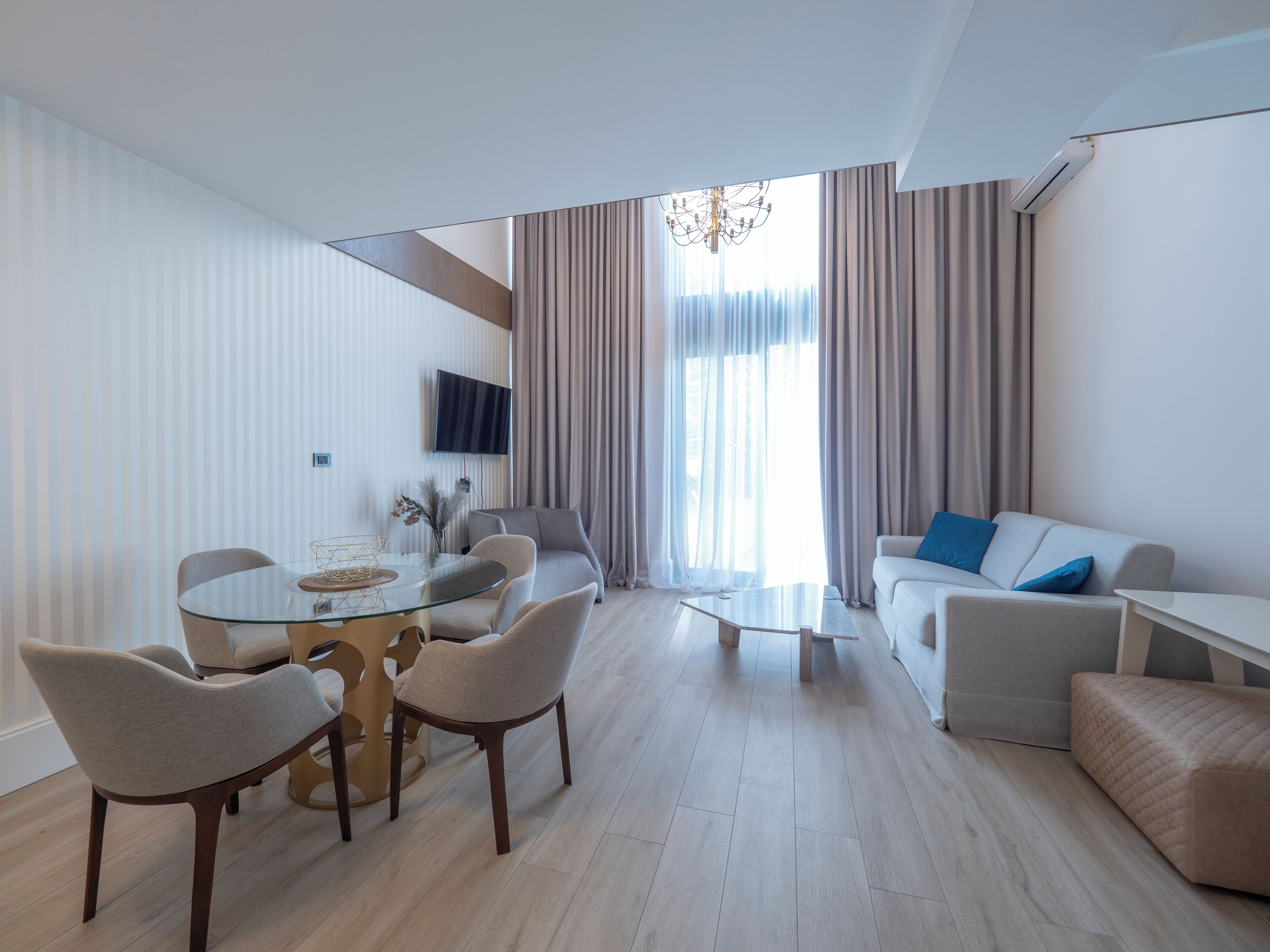 A modern living room with a glass dining table, comfortable seating, a flat-screen TV, and a large window with curtains.