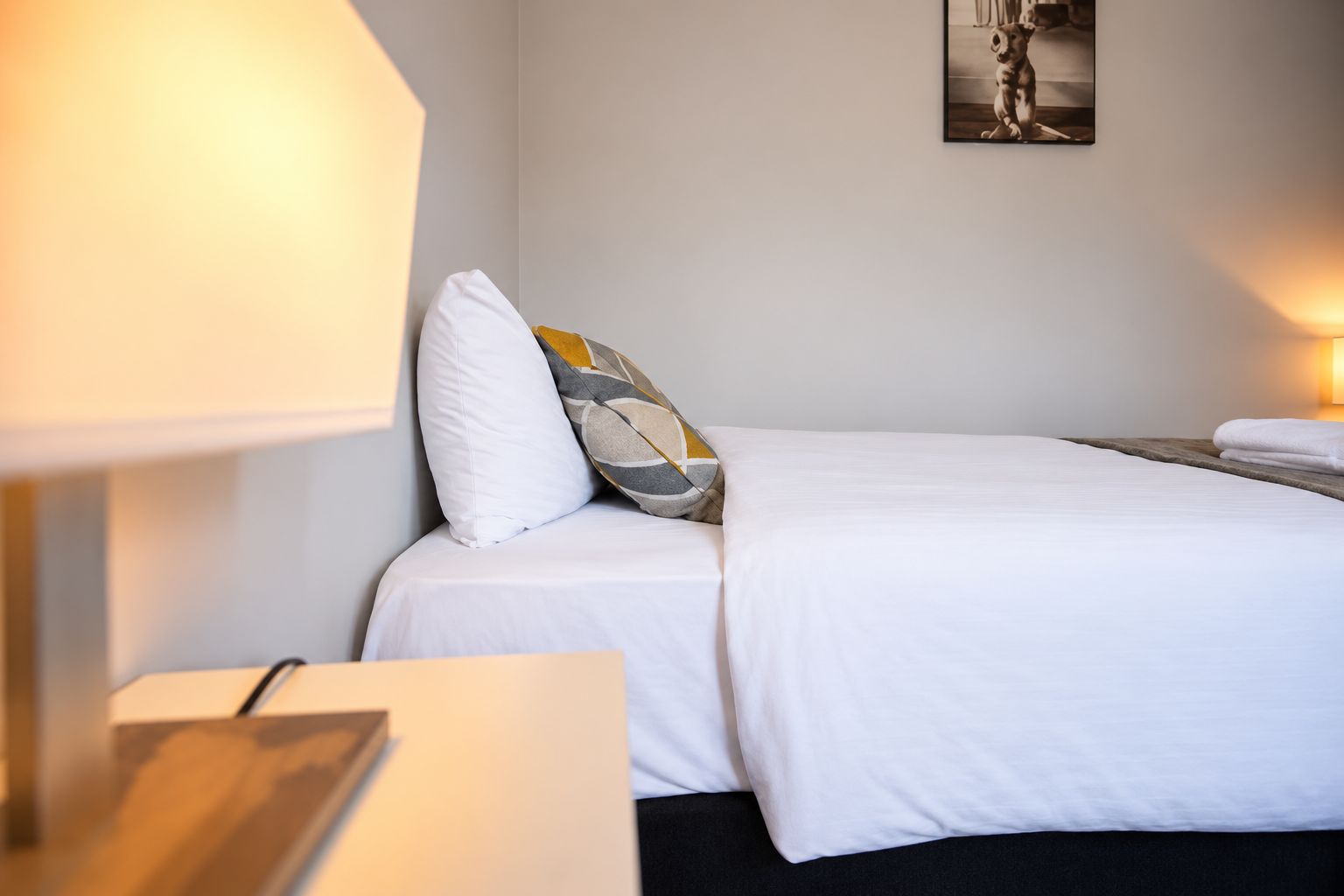 Cama bem arrumada com roupa de cama branca, um travesseiro e mesa de cabeceira com abajur.