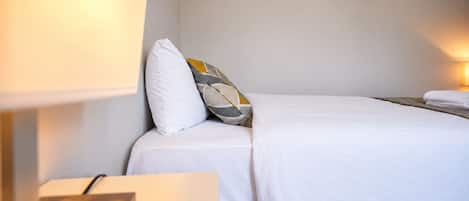 Cama bem arrumada com roupa de cama branca, um travesseiro e mesa de cabeceira com abajur.