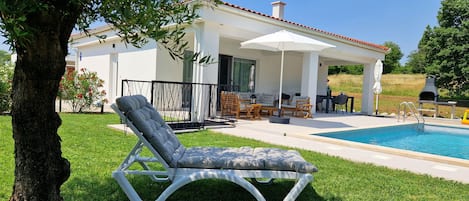 Ett modernt utomhusområde med en pool, en solstol, en täckt uteplats med utemöbler och en grill.
