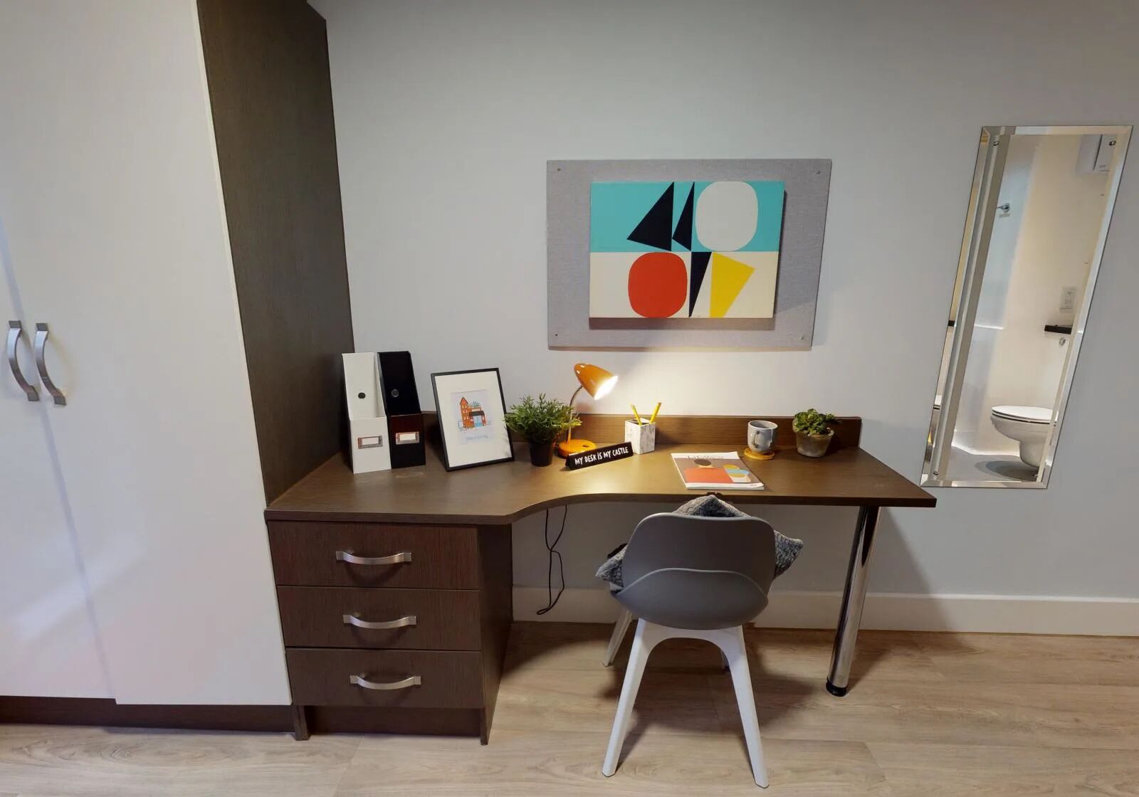 A modern office space with a desk, chair, and a framed picture. There is a potted plant on the desk and a mirror on the wall.