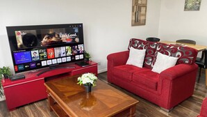 A living room with a flat-screen TV displaying various channels, a red sofa with patterned cushions, a wooden coffee table, and a small plant.