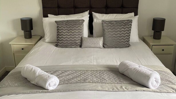 A neatly made bed with white linens, grey patterned pillows, and two rolled towels.