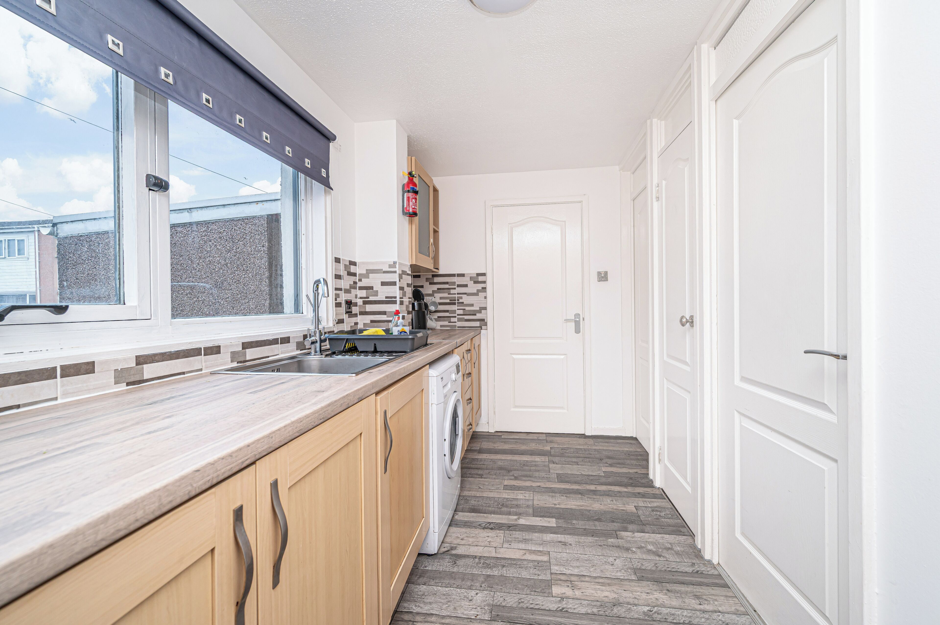 A modern kitchen with a built-in washing machine, a window with blinds, and a door leading to another room.